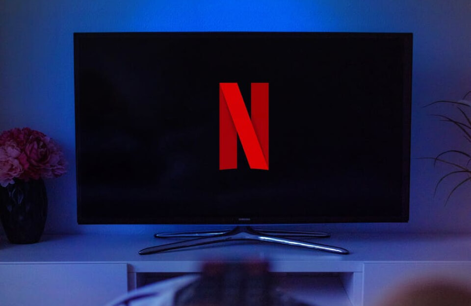 شاشة تلفاز سوداء بإضاءة زرقاء وشعار Netflix الأحمر N في الوسط، مع قاعدة معدنية ولمسات أجواء داكنة في الخلفية